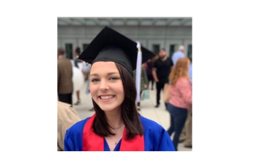 [ai] A young woman in a graduation cap and gown, smiling at the camera. She is wearing a blue gown with a red sash and stands outdoors, with people celebrating in the background.